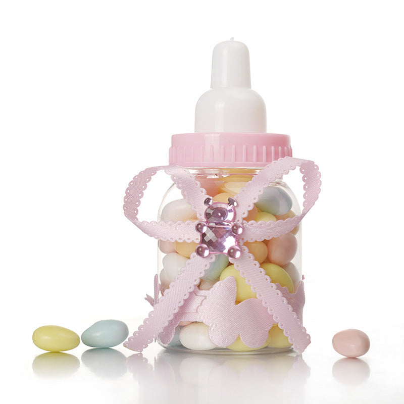 A pink baby bottle container filled with pastel-colored candy, decorated with a pink ribbon and a bear-shaped gem. A few candies are scattered around it on a white surface.
