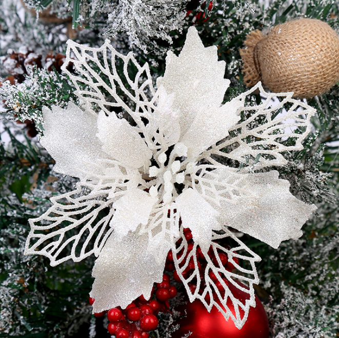 A glittery white poinsettia with delicate, lace-like leaves is nestled in snowy evergreen branches, accompanied by red berries and a burlap ornament.