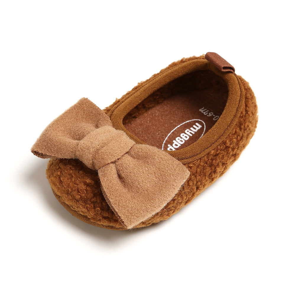 Brown baby shoe with textured fabric, featuring a large, soft bow on the front. The insole displays branding text. The shoe has a plush, cozy appearance.
