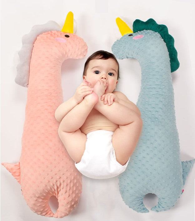 A baby in a diaper lies between two plush dinosaur pillows—one pink with a yellow horn, the other blue, also with a yellow horn. The baby is playfully holding a foot.