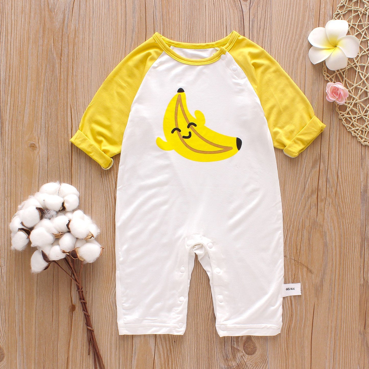Baby romper with yellow banana design on white fabric and yellow sleeves, laid on wooden floor. Cotton plant on the left and flowers on the right as decor.