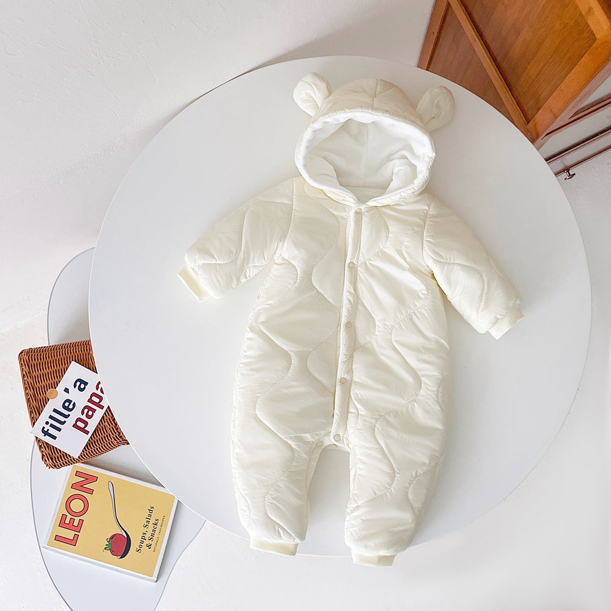 White baby snowsuit with bear ears and button closure, laid on a round table. Nearby are a wicker basket and a cookbook titled "LEON."