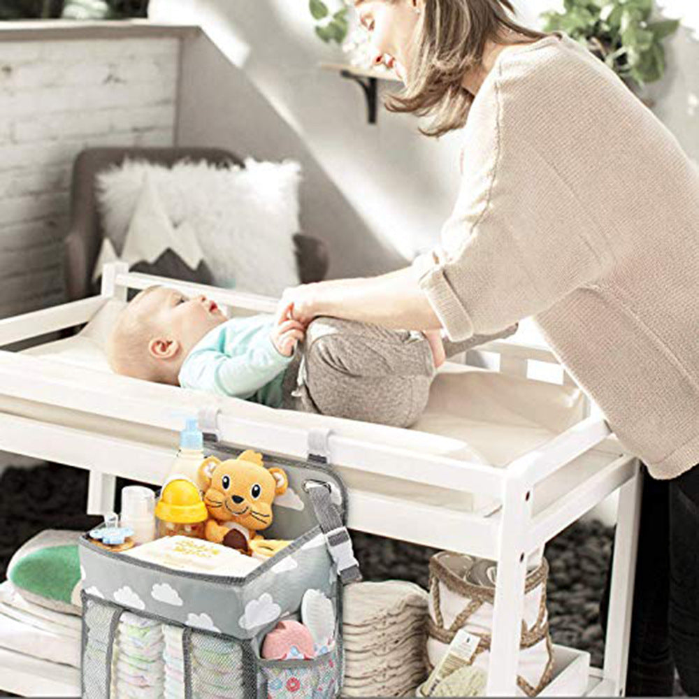 A woman changes a baby on a white changing table. A hanging organizer with baby supplies, including a plush toy and bottles, is attached to the table.