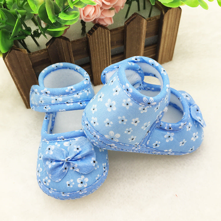 Blue baby shoes with white floral patterns and bows, placed on a white surface. A wooden fence with green and pink flowers is in the background.
