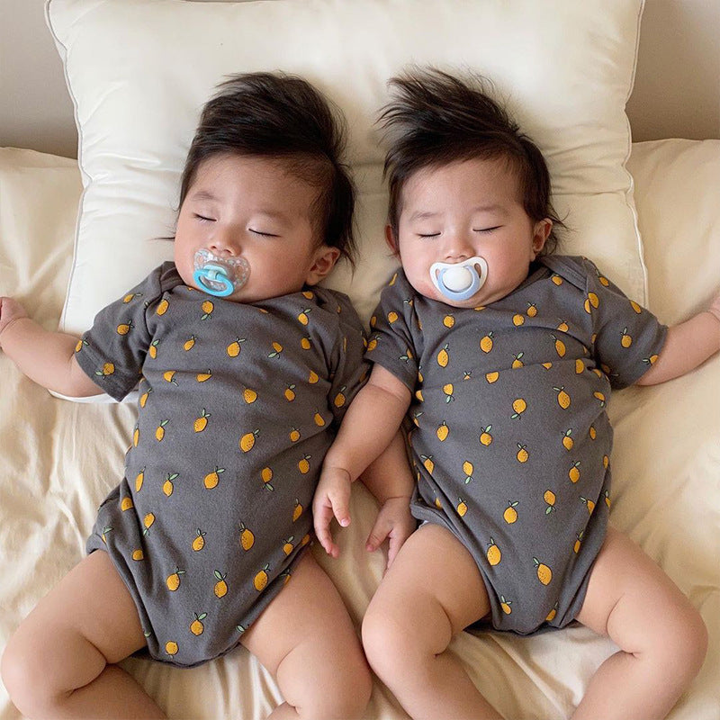 Two sleeping babies, each with a pacifier, wearing matching gray onesies with a small orange fruit pattern. They lie side by side on a white pillow and bed.