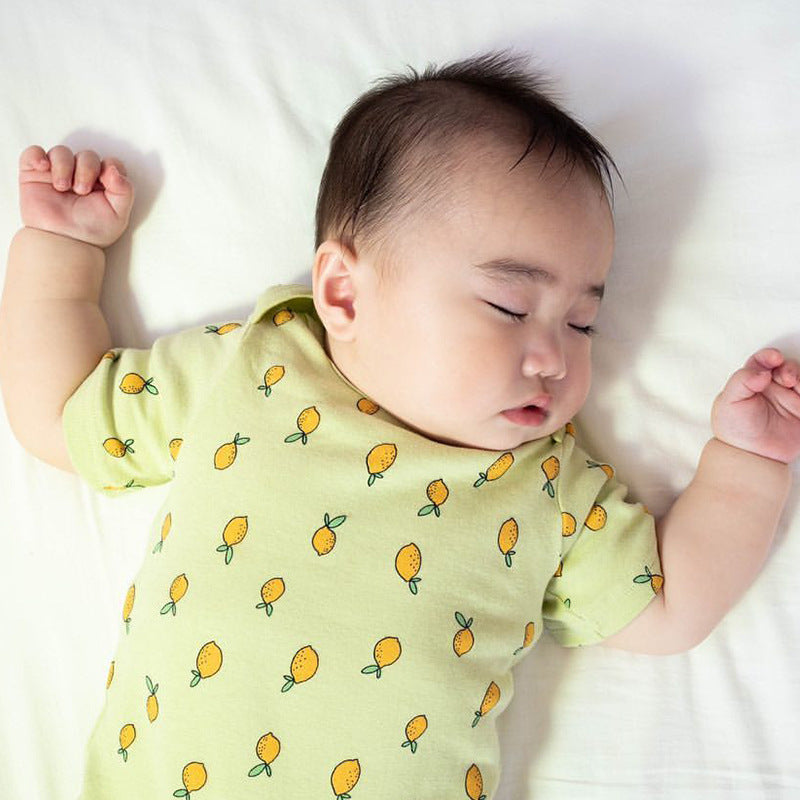 A baby sleeps peacefully on a white sheet, wearing a light green onesie adorned with a pattern of yellow lemons.