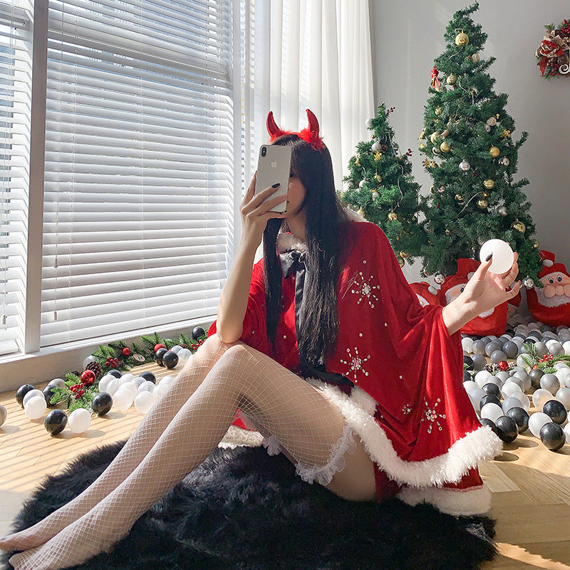 A person in a festive Santa-themed outfit, with red horns, takes a selfie by a decorated Christmas tree. White blinds let in sunlight, highlighting ornaments scattered on the floor.