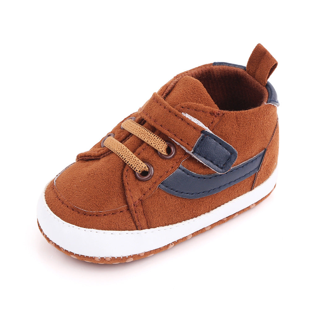 Brown suede baby shoe with tan laces, navy side stripe, and white sole. Features a Velcro strap and a small pull tab at the heel for easy wearing.