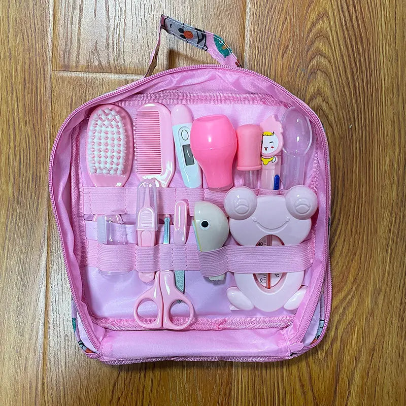 Pink baby care kit on wooden floor, containing essentials like a hairbrush, comb, thermometer, scissors, nail clipper, suction bulb, and a frog-shaped thermometer protector.