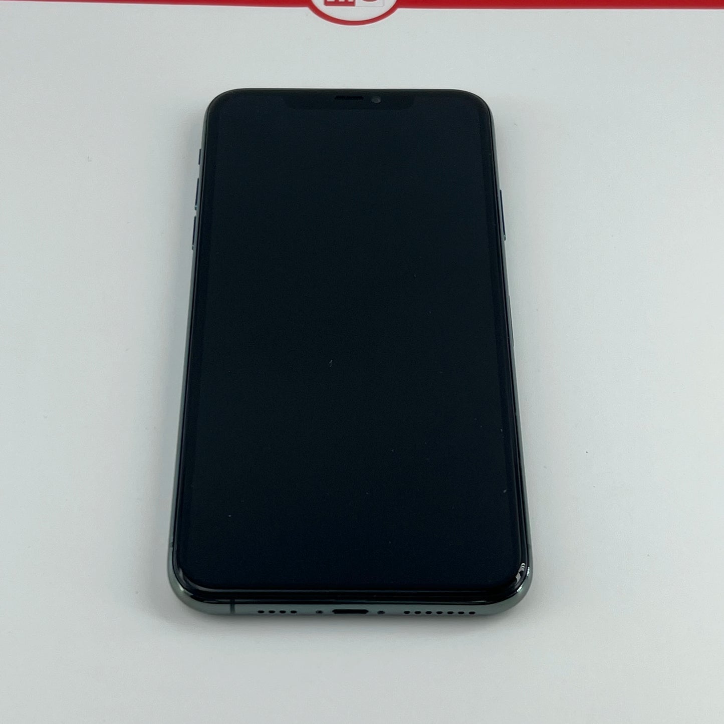 A black smartphone with a blank screen lies on a white surface, featuring a notch at the top and visible side buttons. The red and white design is partially visible above.