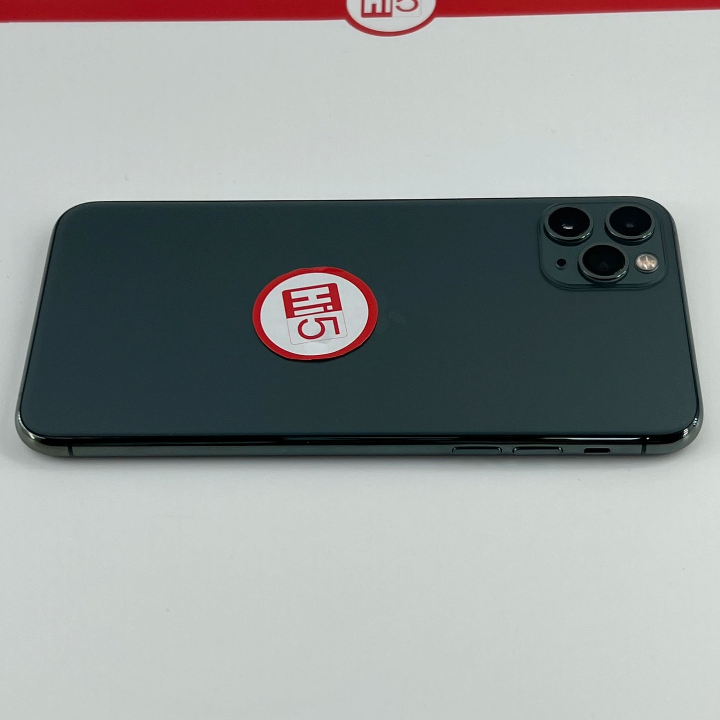 A smartphone with a dark, sleek back lies on a surface. A red and white sticker with "Hi5" is placed near the triple camera lens. A similar logo appears on the backdrop.
