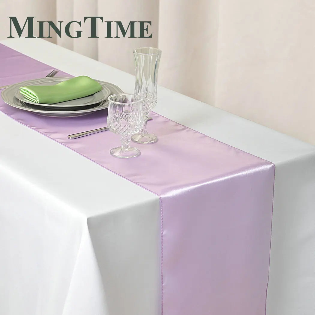 Elegant table setting with a lavender satin runner on a white tablecloth, featuring crystal glasses, a stack of plates with a green napkin, and silver cutlery.