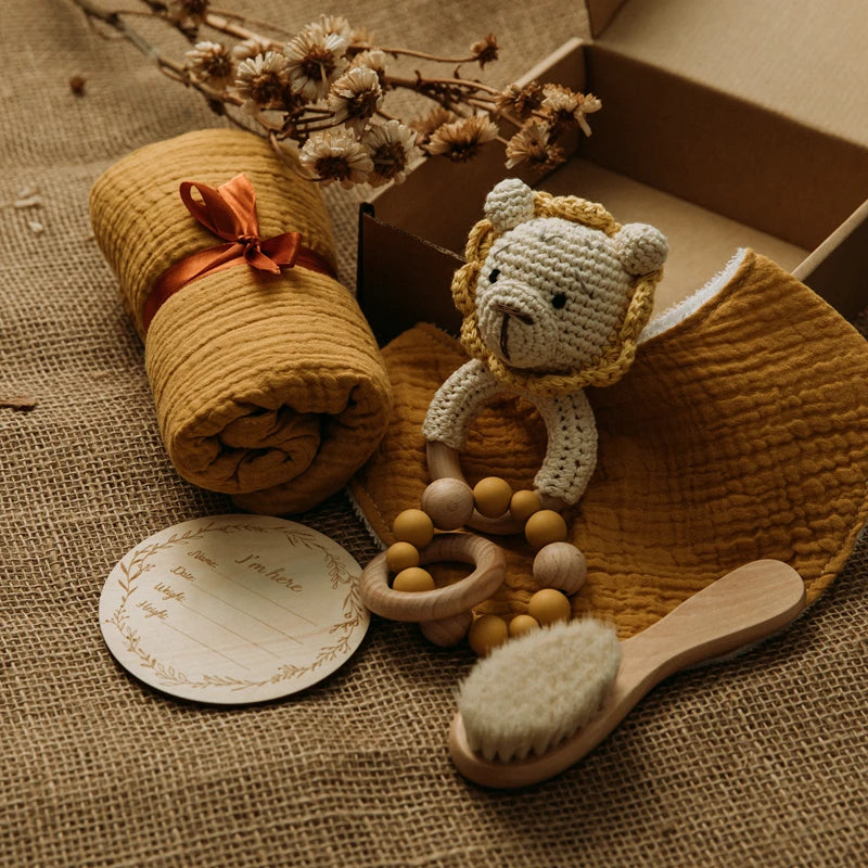 A cozy baby gift set on burlap includes a mustard yellow muslin blanket, lion crochet rattle, wooden brush, teether, and a round milestone card with flowers in the background.
