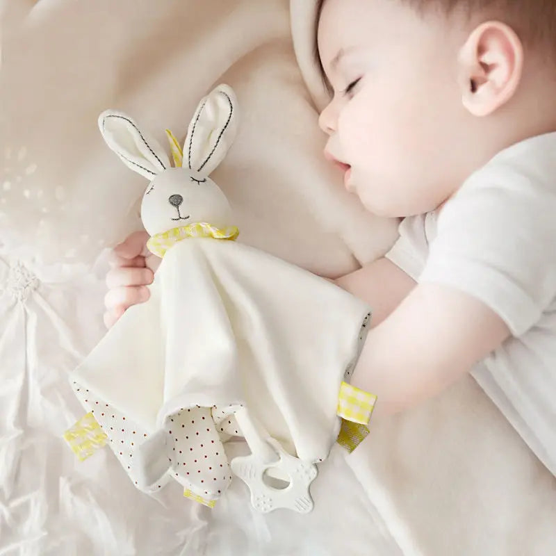 Sleeping baby in a white onesie cuddling a soft bunny toy. The bunny has a fabric body and crinkly ears, with yellow and polka dot accents, lying on a cream bedspread.