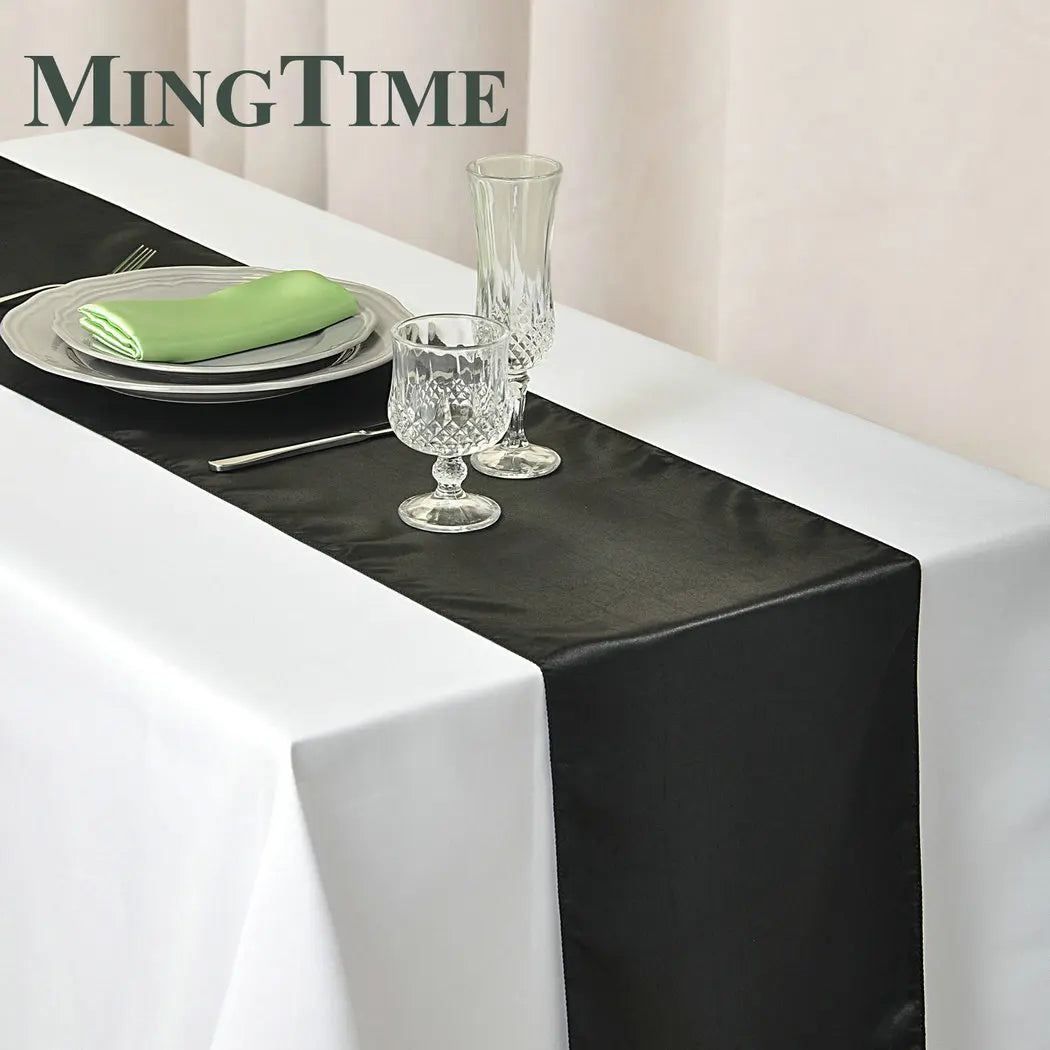 Elegant table setting with a white tablecloth and black runner. Includes crystal glasses, a green napkin on stacked plates, and cutlery, under soft lighting.