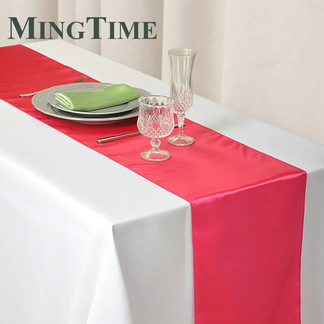 Elegant table setting with a red satin runner on a white cloth, featuring stacked plates with a green napkin, silverware, and two crystal glasses.