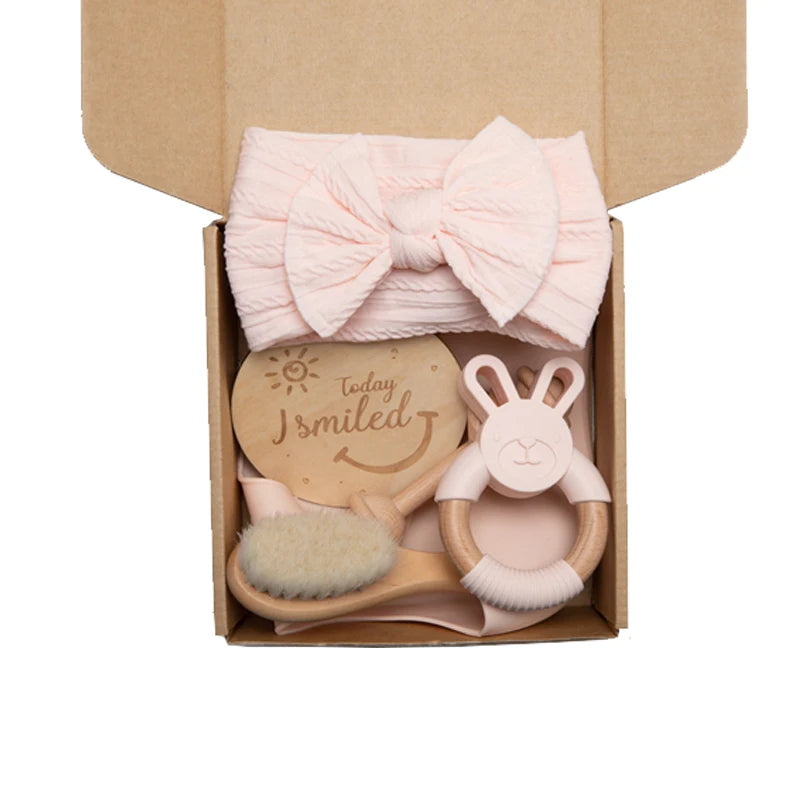 A gift box containing a pink headband with a bow, a wooden brush, a bunny rattle, and a sign reading "Today I smiled" with a sun and smiley face.