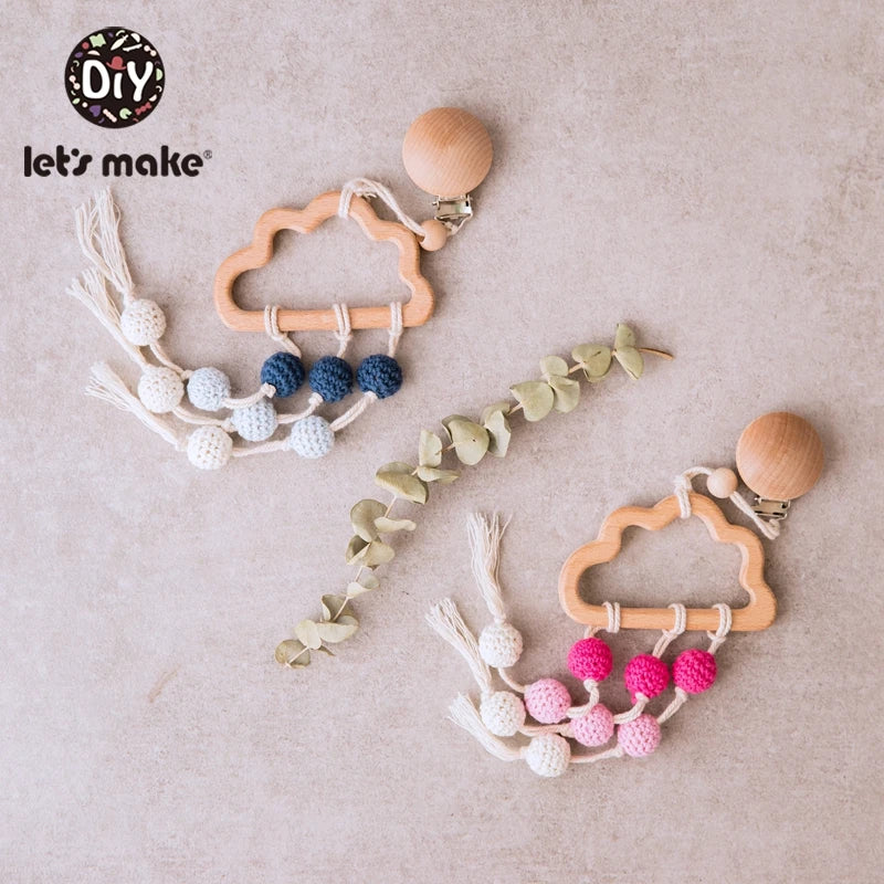 Two cloud-shaped wooden baby teethers with crochet beads in white, blue, and pink. Each has a clip and fringed tassels. Decorative eucalyptus leaves in between.