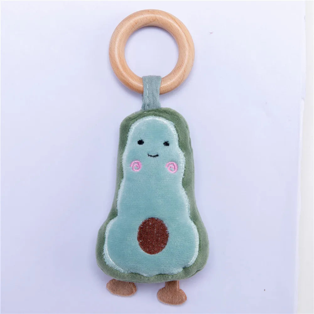 Soft avocado-shaped plush toy with a smiley face and rosy cheeks, attached to a wooden ring. The toy has felt feet and a brown embroidered seed on its belly.