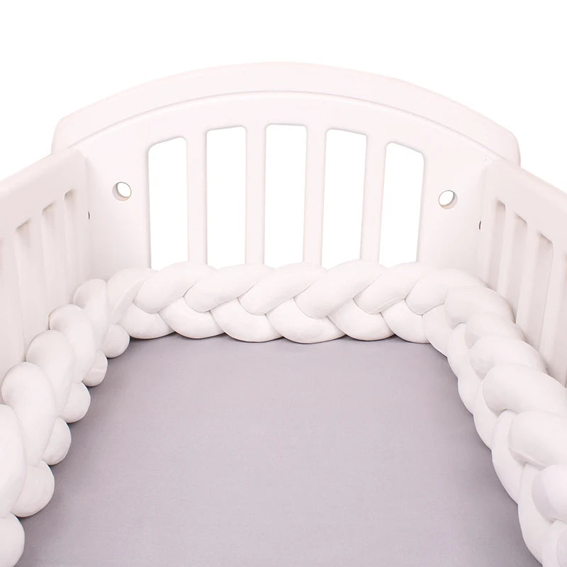 White wooden baby crib with a braided bumper and a soft gray mattress. The bumper is thick and cushioned, running along the inside perimeter for added safety.