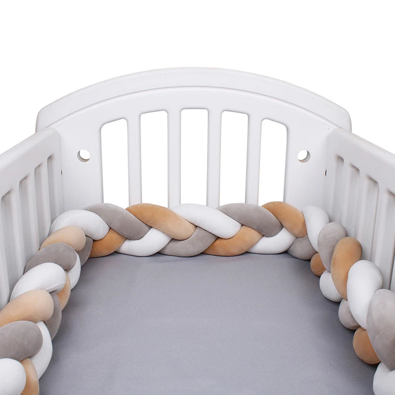 White crib with a gray mattress, featuring a braided bumper in soft shades of beige, white, and gray around the interior for added safety and style.