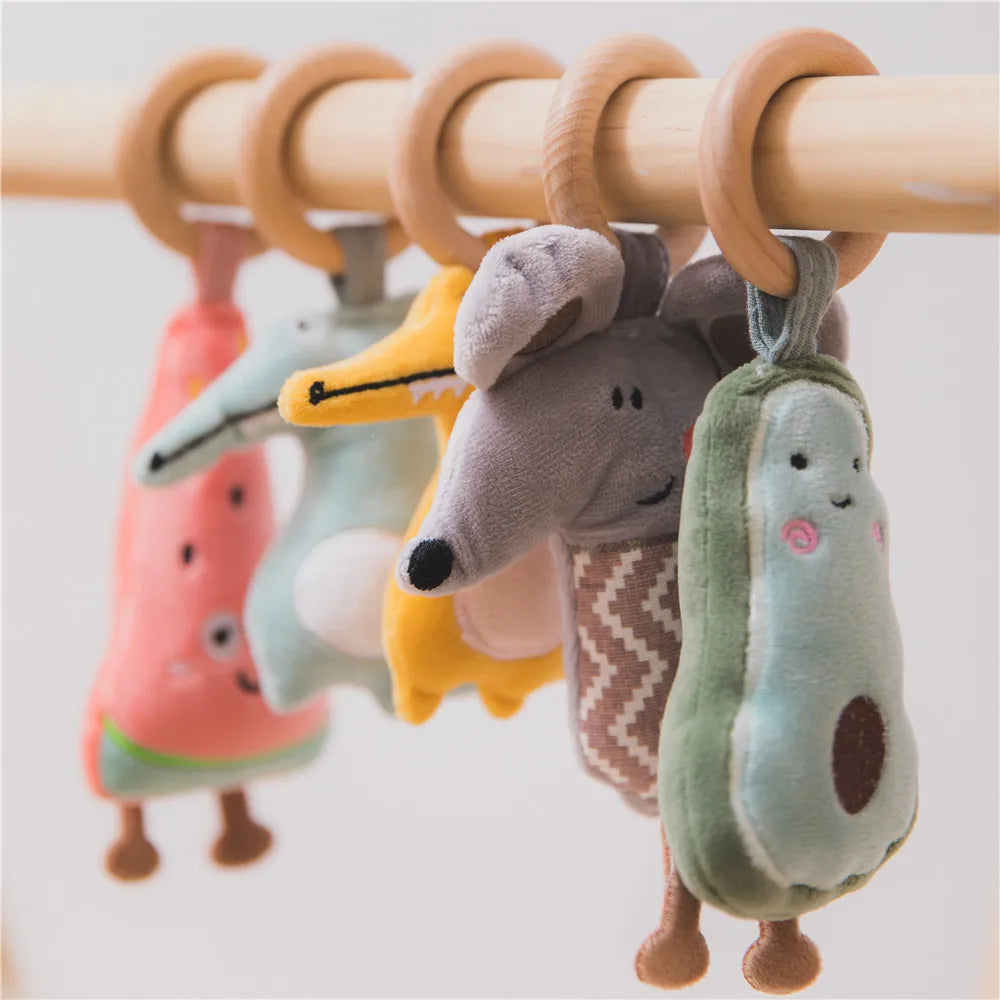 Plush toys resembling various animals and objects, like a watermelon and avocado, hanging from wooden rings on a rail. The toys are colorful and cartoonish.