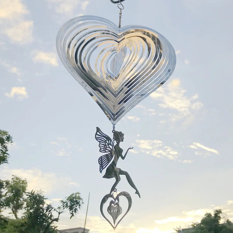 A metallic heart-shaped wind spinner with a dangling fairy silhouette and a smaller heart inscribed with "love" hangs against a cloudy sky with tree silhouettes below.
