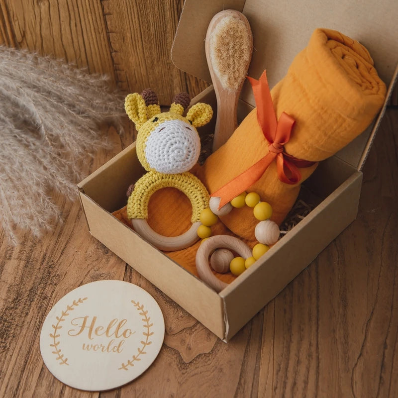 Gift box with an orange swaddle, wooden brush, yellow beaded teether, and a crochet giraffe ring on a wooden table. A “Hello world” plaque lies beside the box.