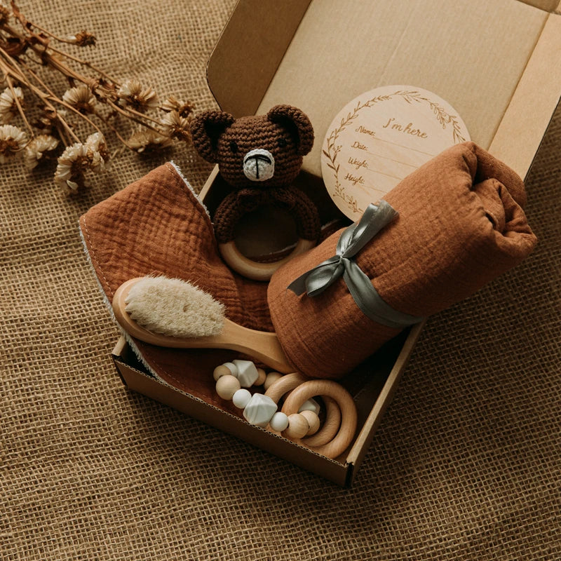A gift box on burlap contains a brown baby blanket, a wooden hairbrush, a crocheted teddy bear rattle, wooden teething rings, and a round wooden birth announcement plaque.