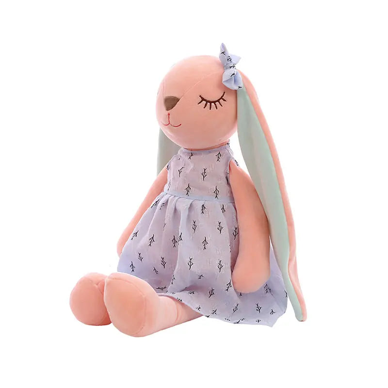 A soft, plush bunny toy with long ears, dressed in a light purple dress with small tree prints, and a matching bow on its head, sitting with eyes closed.