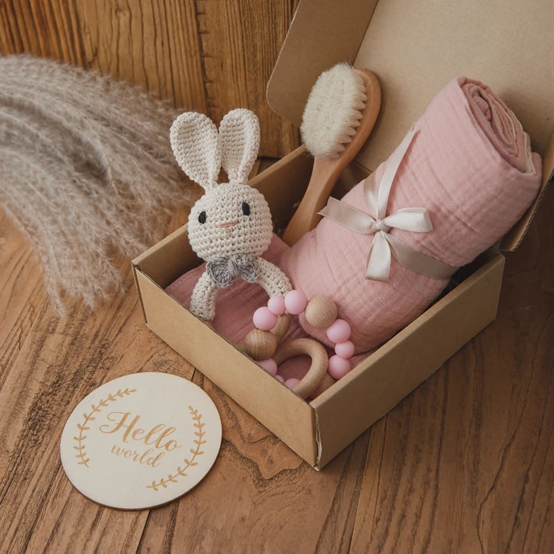 A gift box with a crocheted bunny, pink swaddle, wooden brush, and pink beaded teether. Nearby, a round wooden plaque reads "Hello world" on a rustic wooden surface.