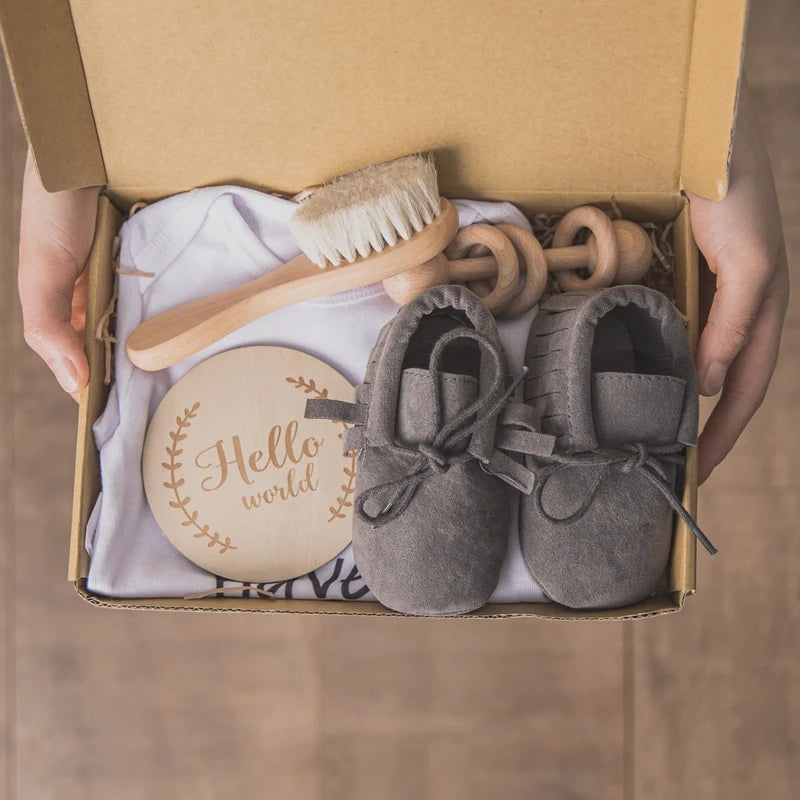 Gift box with baby essentials: gray booties, a wooden brush, a rattle, and a round plaque reading "Hello world." They are placed on a fabric inside the box.