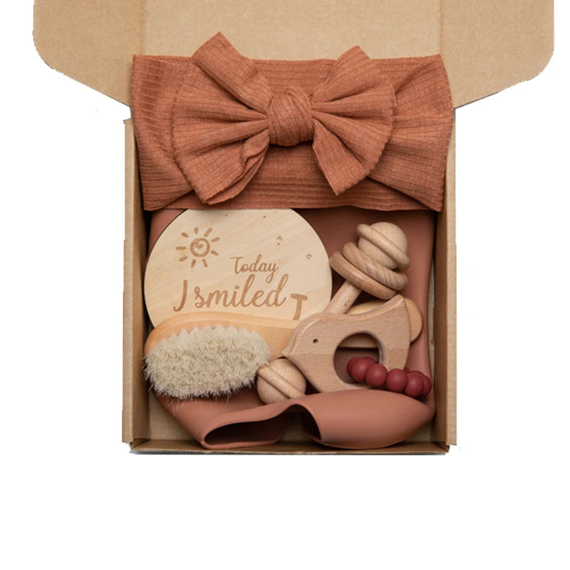 Gift box with a large brown bow, wooden "Today I smiled" plaque, soft baby brush, wooden teether shaped like an elephant, and teether beads, set against a soft brown cloth.