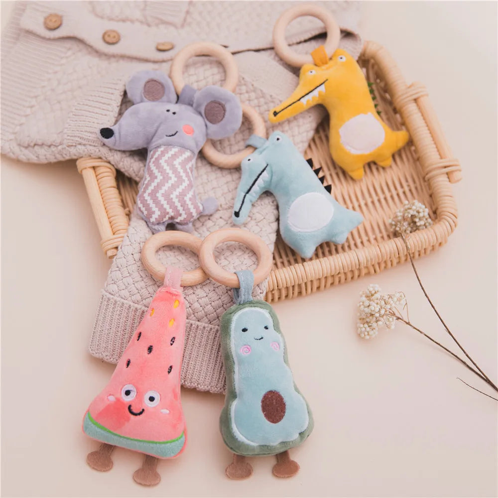 Plush animal and fruit rattles with wooden rings in a basket on a beige surface. Includes a mouse, alligator, watermelon, and avocado designs, with a knitted sweater underneath.