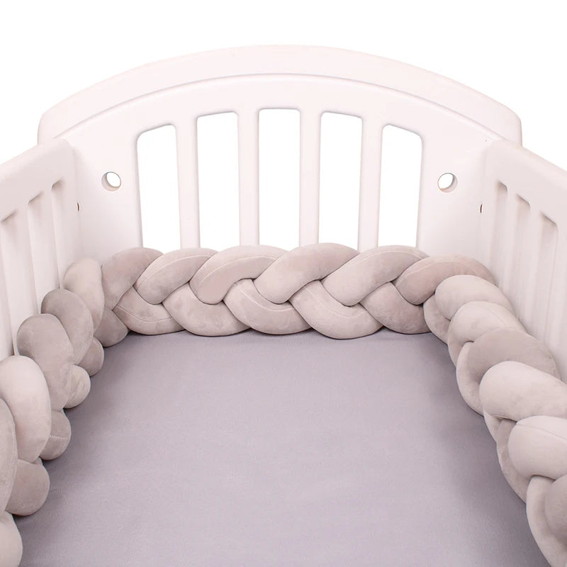 White crib with a soft, braided grey bumper lining the inside. The mattress is covered with a matching grey sheet, creating a cozy and safe sleeping space for a baby.