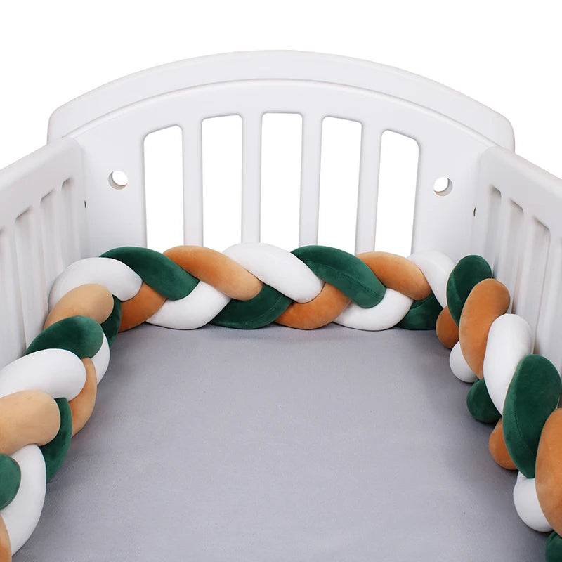 White baby crib with a braided bumper in green, white, and peach colors lining the inside edges. The mattress is covered with a smooth gray sheet.