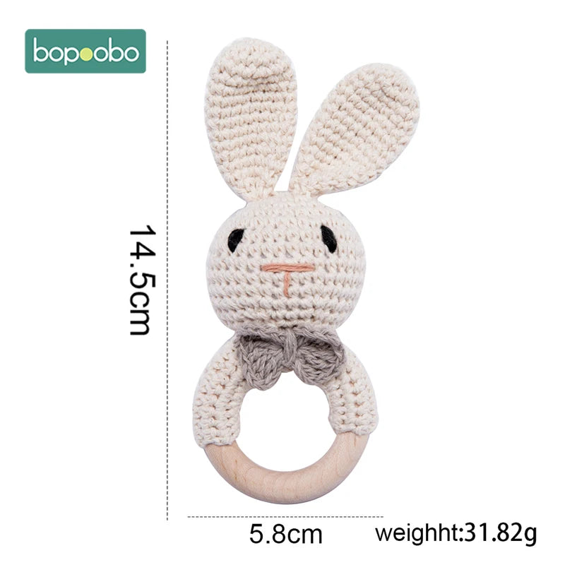 Crocheted bunny rattle with elongated ears, wooden ring, and a gray bow. Dimensions: 14.5cm height, 5.8cm width. Weight: 31.82g. Brand logo at the top left.