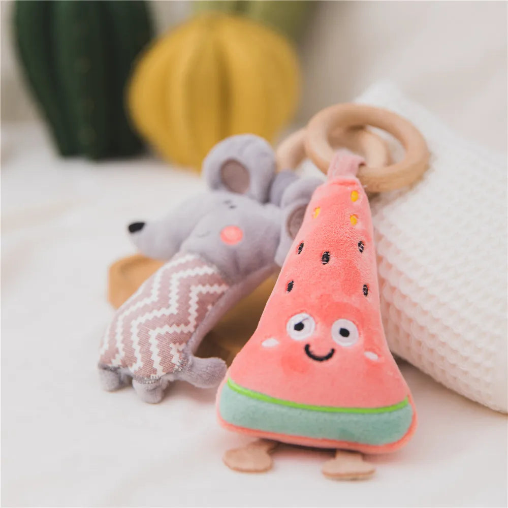Plush toys on a bed: a smiling watermelon slice with legs and a gray mouse wearing a zigzag-patterned outfit, with wooden rings attached, blurred background.