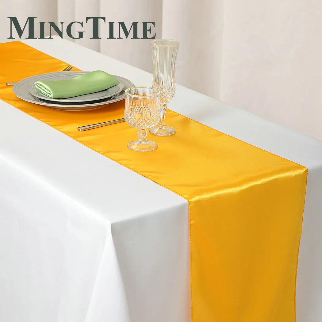 White table with a yellow satin runner, set with a green napkin on a gray plate, two crystal glasses, and cutlery. The background has soft drapery. Text reads "MINGTIME".