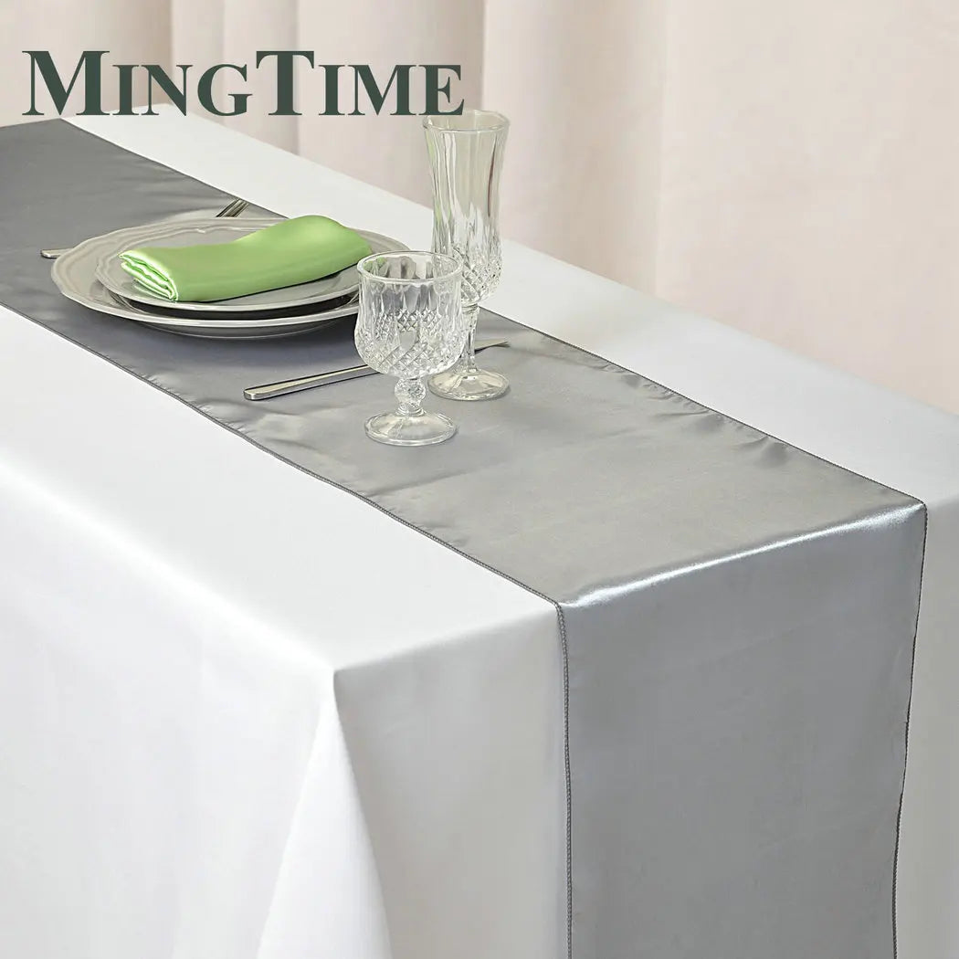 Elegant table setting with a gray satin table runner on a white cloth, featuring stacked plates, a green napkin, silverware, and crystal glassware in a minimalist arrangement.