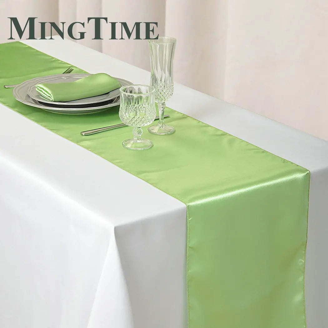 Elegant table setting with a white tablecloth and a light green satin runner. Features a plate, folded napkin, fork, glassware, and flutes. "MINGTIME" logo in the corner.