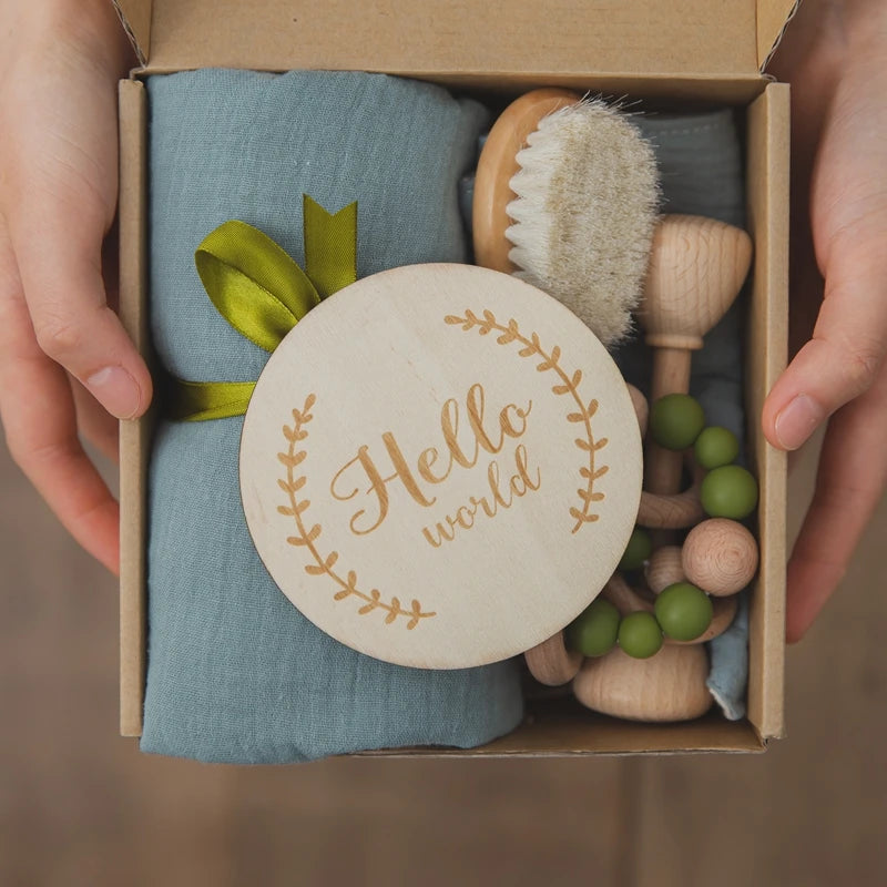 Hands holding a gift box containing a "Hello world" wooden sign, a baby brush, a teether with green beads, and a blue cloth tied with an olive green ribbon.