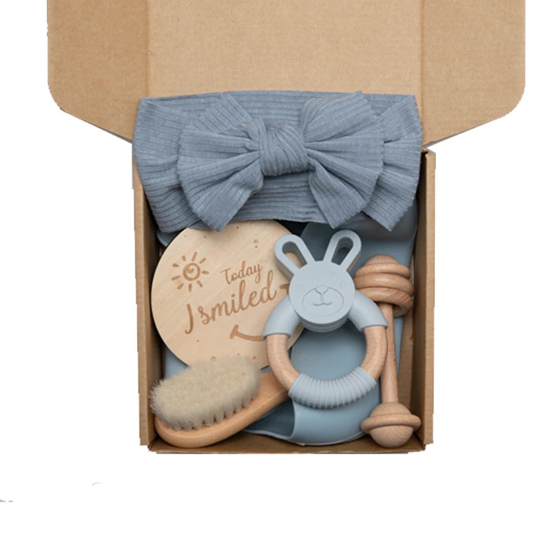 A cardboard box containing a gray bow headband, a wooden disc with "Today I smiled," a bunny-shaped teether, baby hairbrush, and a knotted rope toy.