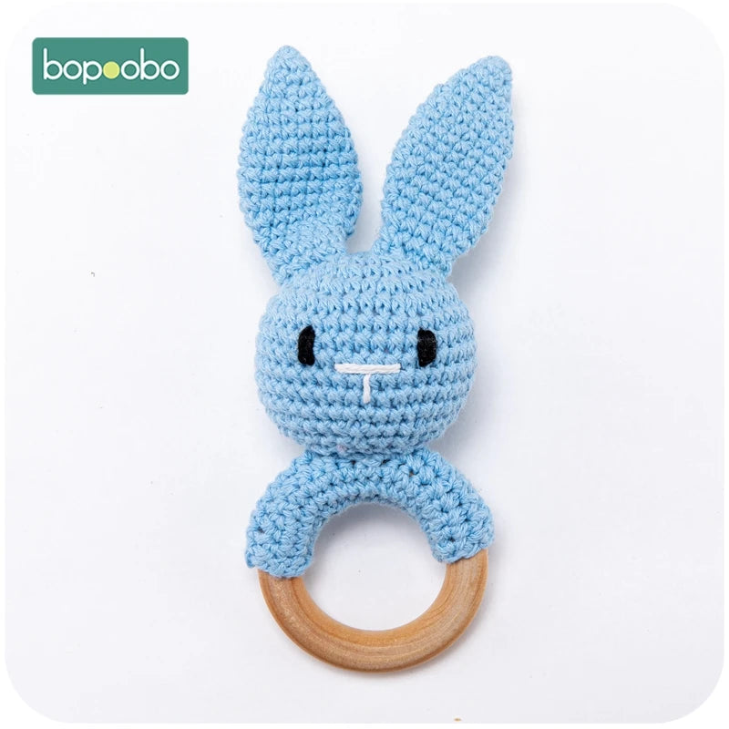 Crocheted blue bunny with long ears and black facial features, attached to a wooden ring, centered on a white background. Bopoobo logo in the corner.