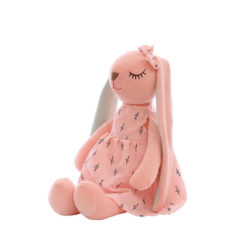 Pink plush bunny with long ears wearing a matching dress decorated with small black patterns. The bunny has closed eyes and a small bow on its head, sitting upright.