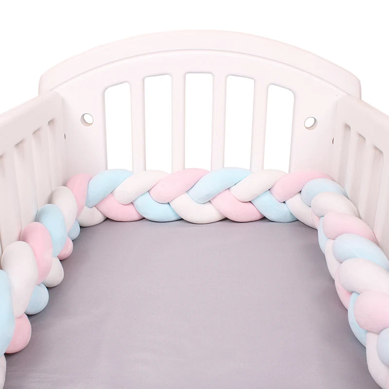 White crib with a grey mattress and a soft, braided bumper in pastel pink, blue, and white colors lining the interior.