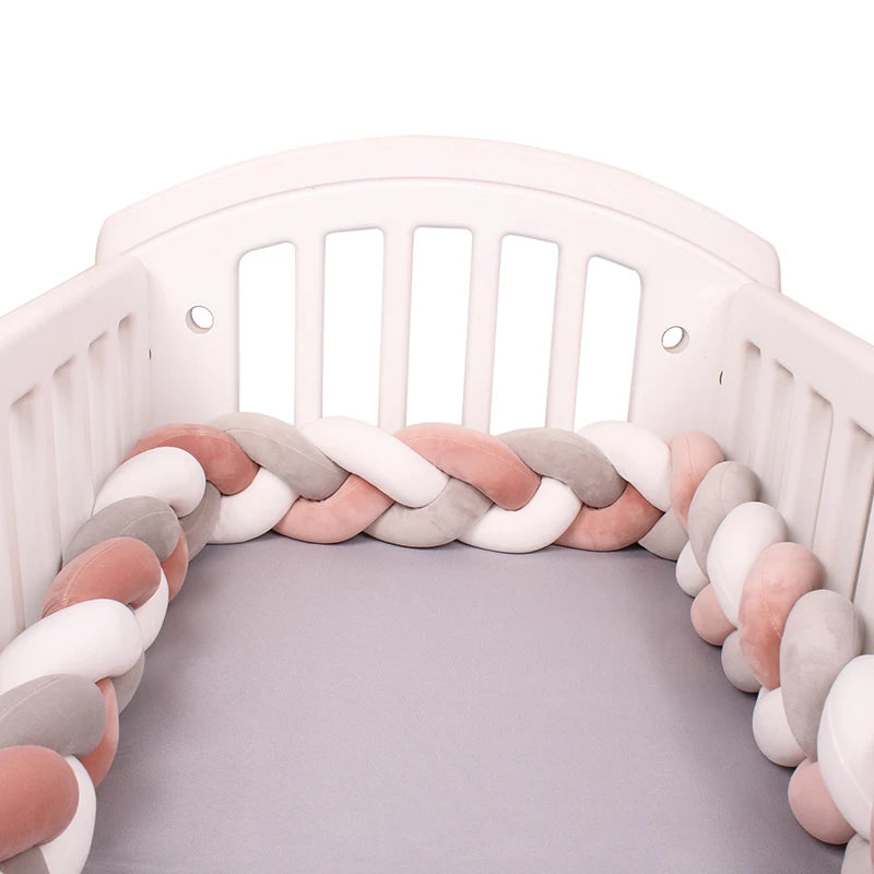 A white crib with a braided bumper in soft pastel colors (pink, gray, white) lines the inside. The mattress is covered with a light gray sheet.