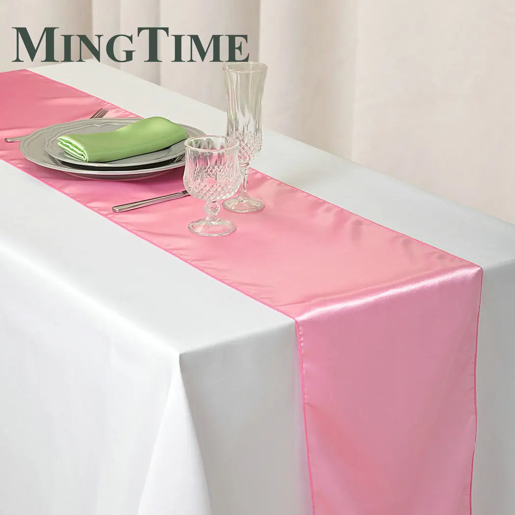 Elegant dining setup with a pink satin table runner over a white tablecloth, featuring stacked plates with a green napkin, crystal glassware, and cutlery.