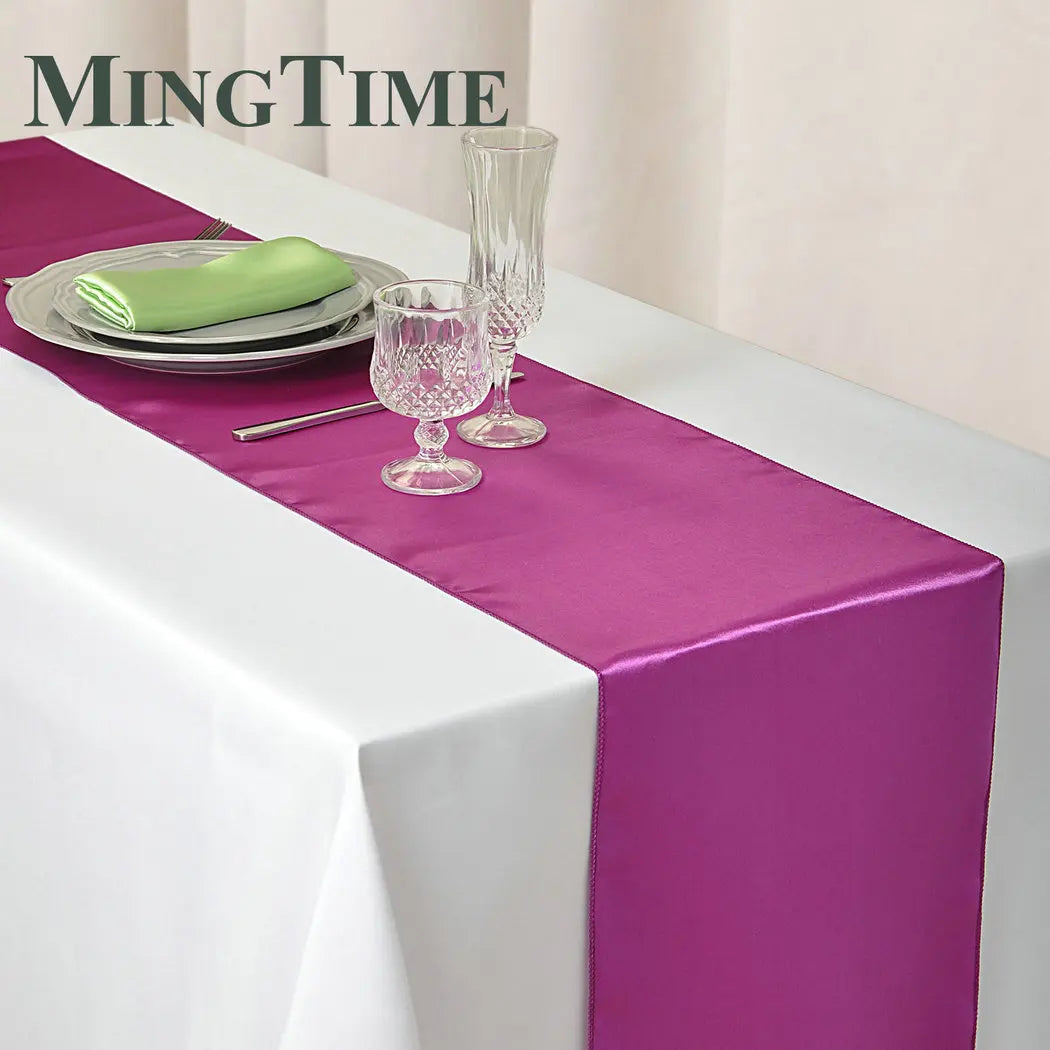 Elegant dining table with a white tablecloth and a vibrant purple runner. It is set with crystal glassware, silver cutlery, and stacked plates with a green napkin.