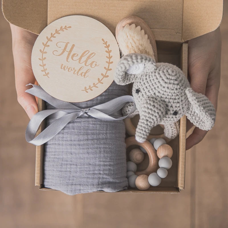 Gift box with a knitted elephant toy, gray cloth wrapped in a ribbon, wooden brush, teething ring, and a “Hello World” sign on top.